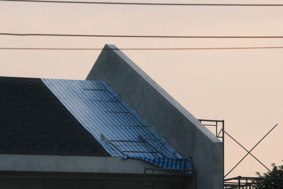 Unfinished concrete gable with a blue tarp roof, power lines and an X-braced scaffold against a peach sky
