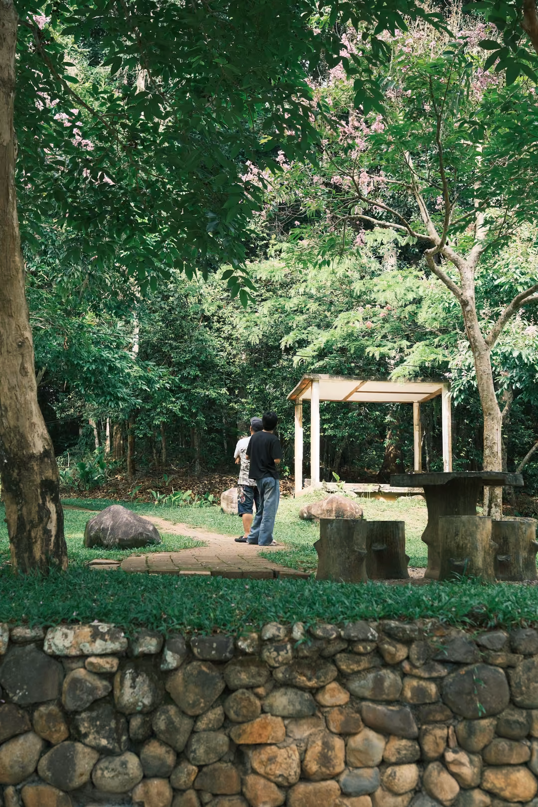 Two people standing by a small white shelter in a shaded garden, above a low stone wall