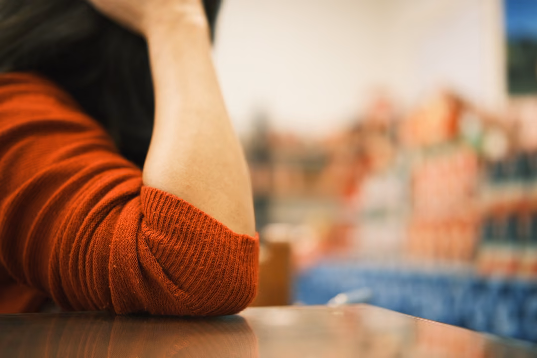Close view of a forearm in a rust-red sleeve resting on a glossy table, the shop blurred behind