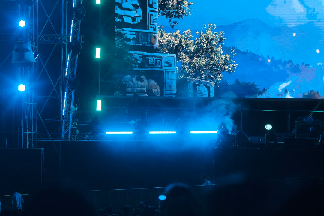 Stage side with green lights and steel scaffolding, a screen showing a stone ruin and a tree under blue sky.