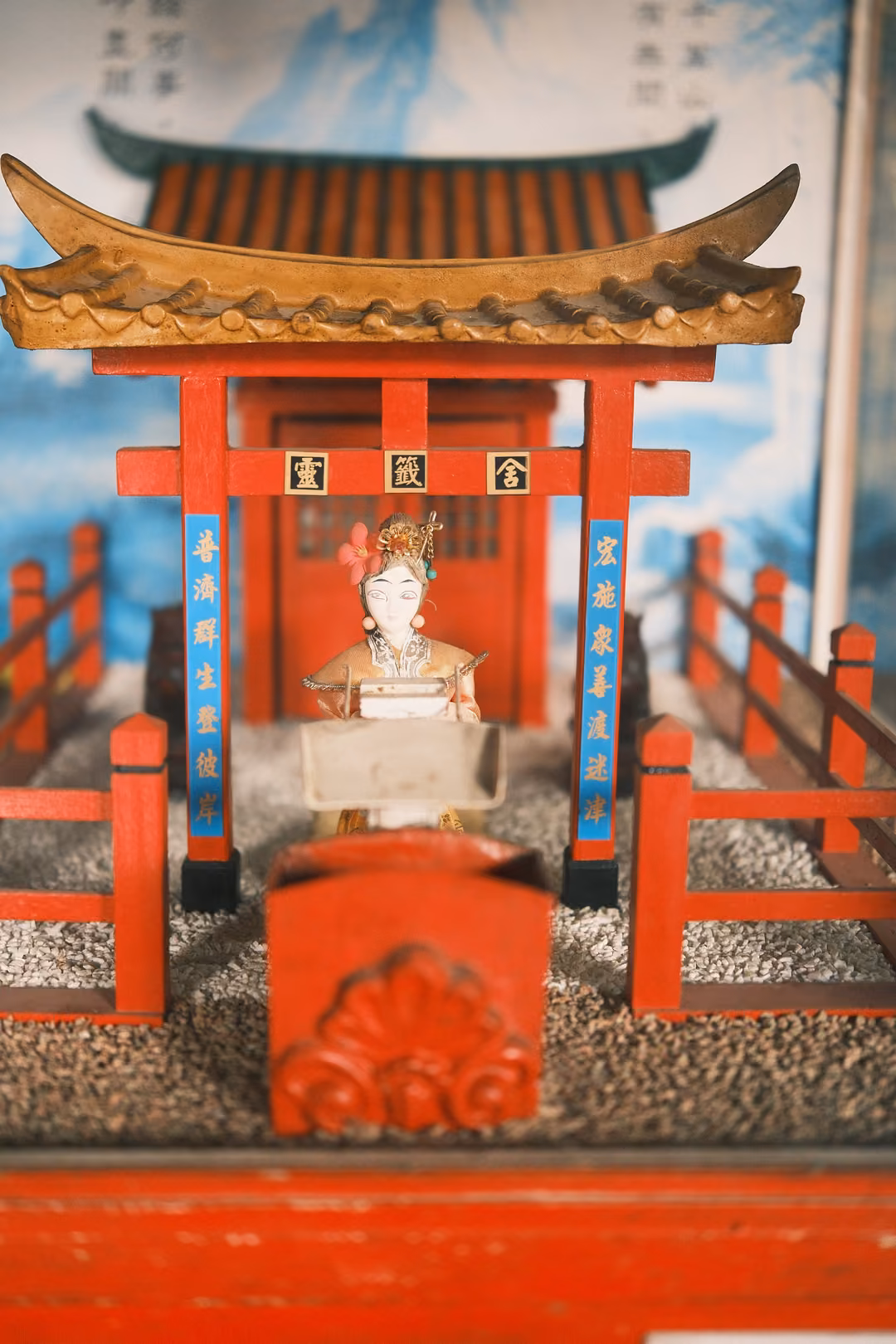 Miniature red shrine with a curved golden roof and a small crowned figure, painted sky backdrop behind