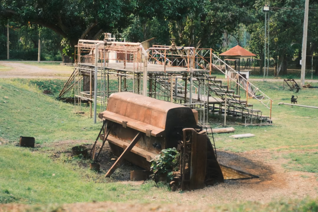 A large rusted metal machine over a dirt pit on a grassy slope, metal scaffolding and stairs behind.