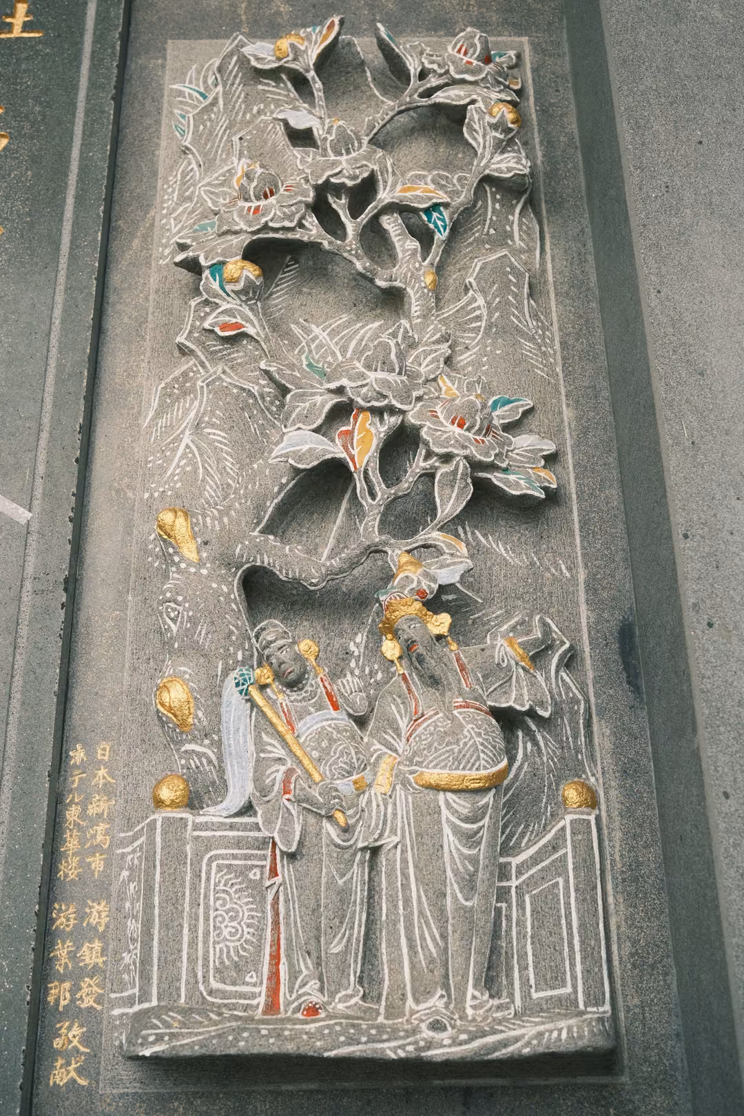 Tall stone relief panel of flowering branches above two robed figures, with gold inscriptions at the base