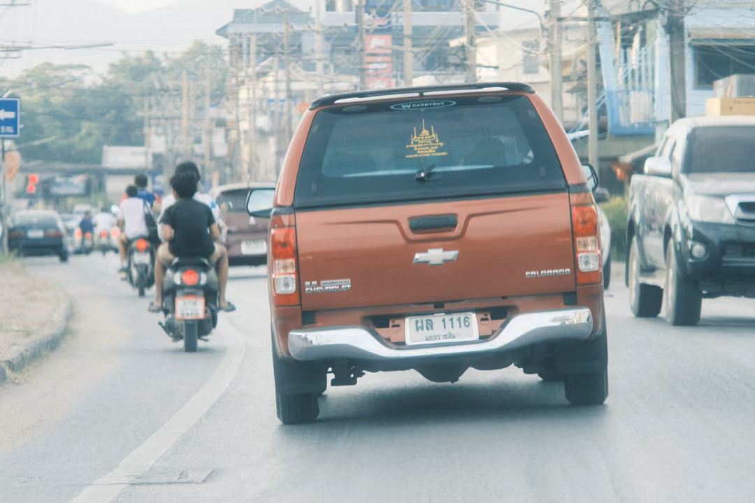 An orange-red Chevrolet pickup drives down a town road behind motorbike riders, shops and power lines alongside.