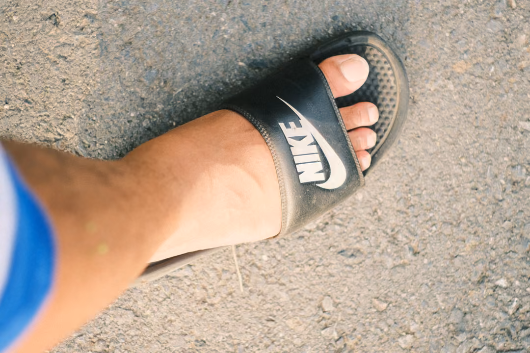 Close view of a foot in a black slide sandal with white logo on grey asphalt