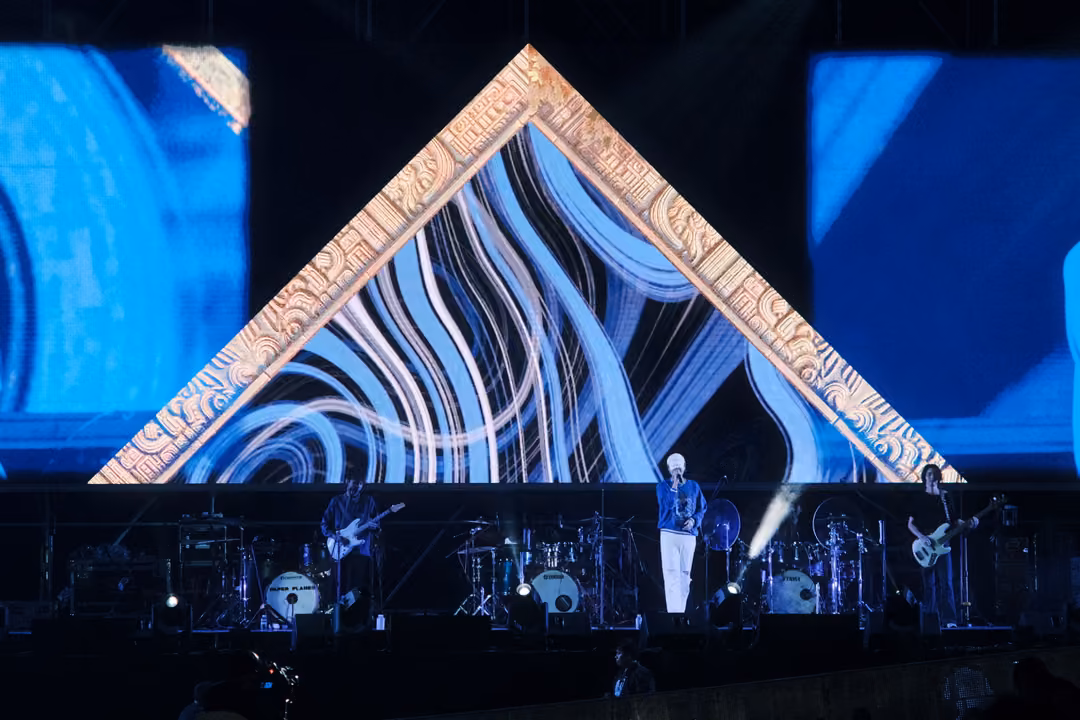Singer in white trousers at centre stage beneath a gold triangular frame filled with white and blue swirls.