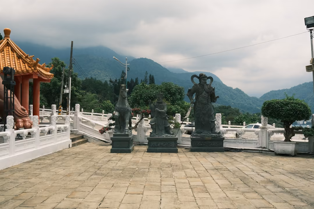 Temple courtyard with dark stone statues on plinths, a red and gold pavilion at left, cloud-covered mountains