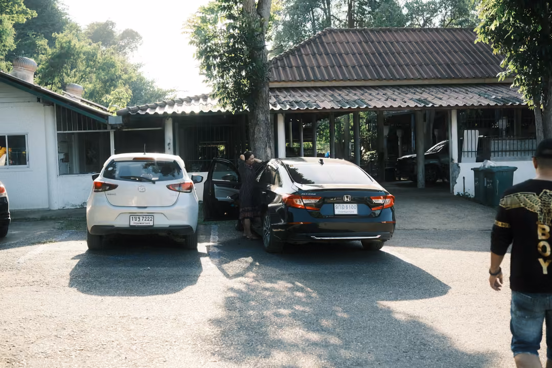 Parking area with a white hatchback and a dark sedan near a tiled-roof building, a man walking in at right