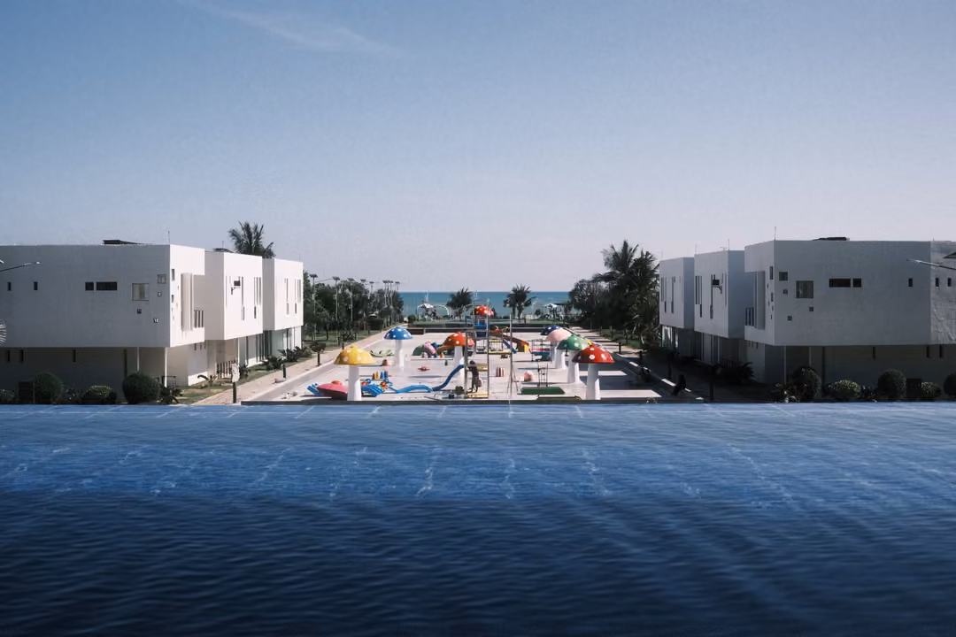 Infinity pool surface in the foreground leading to white villas, a colourful splash park and a strip of ocean