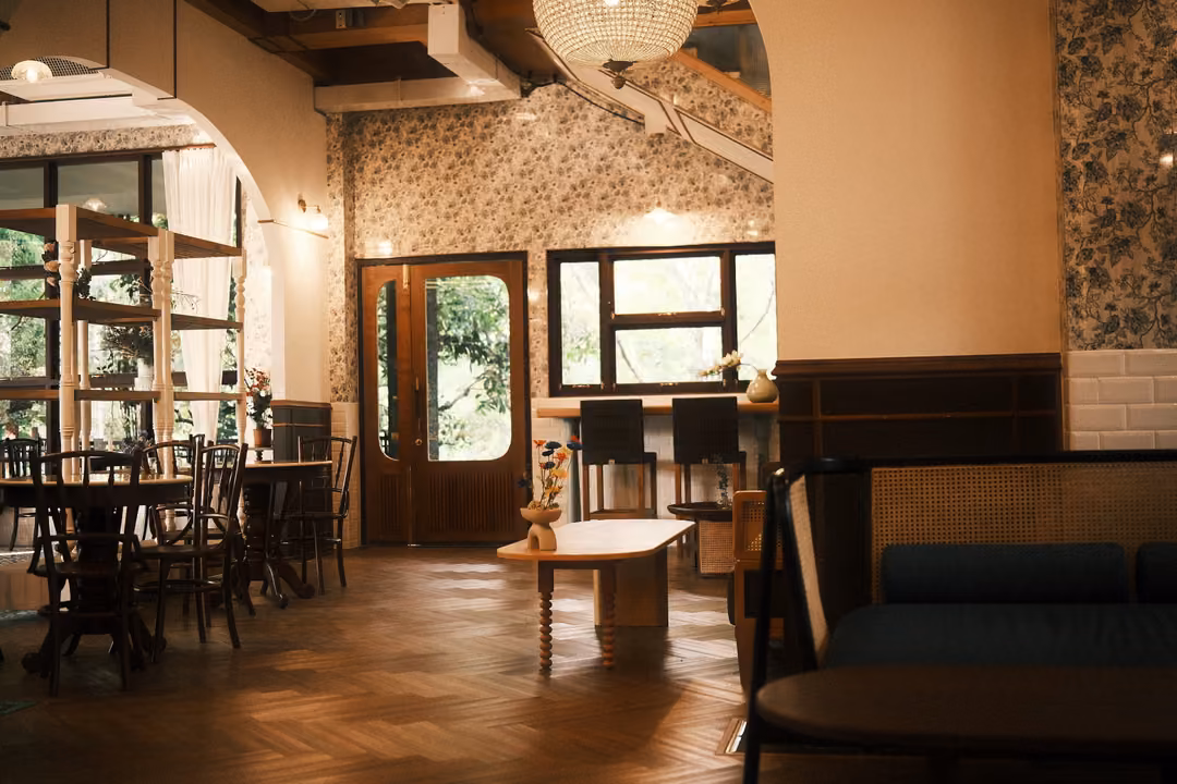 Wide view of a café room with floral wallpaper, an arched doorway, wooden tables and a dark sofa