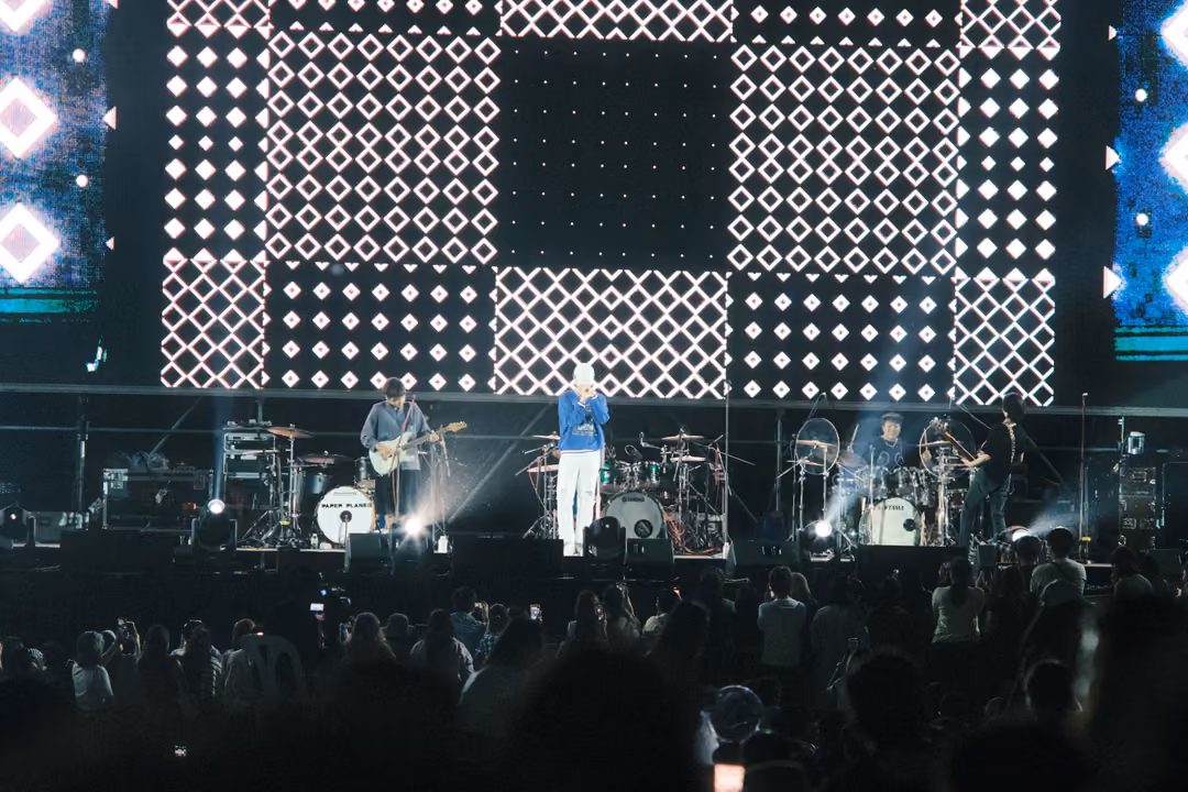 Band on stage below a large screen of white diamond tiles, the crowd in the foreground, a phone screen glowing.
