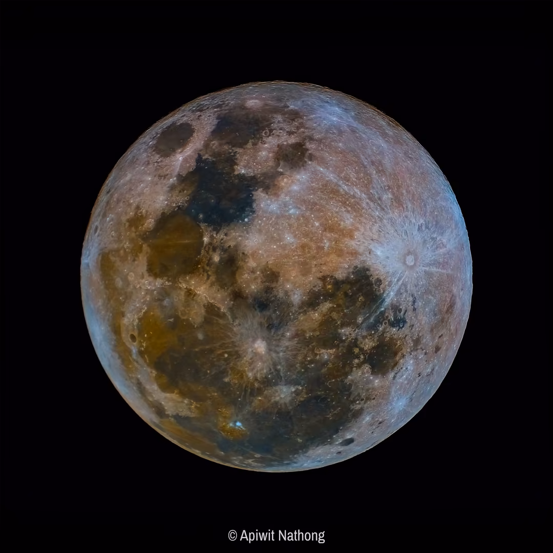 A full moon in vivid false colour, blue and gold tones across its surface, bright ray craters