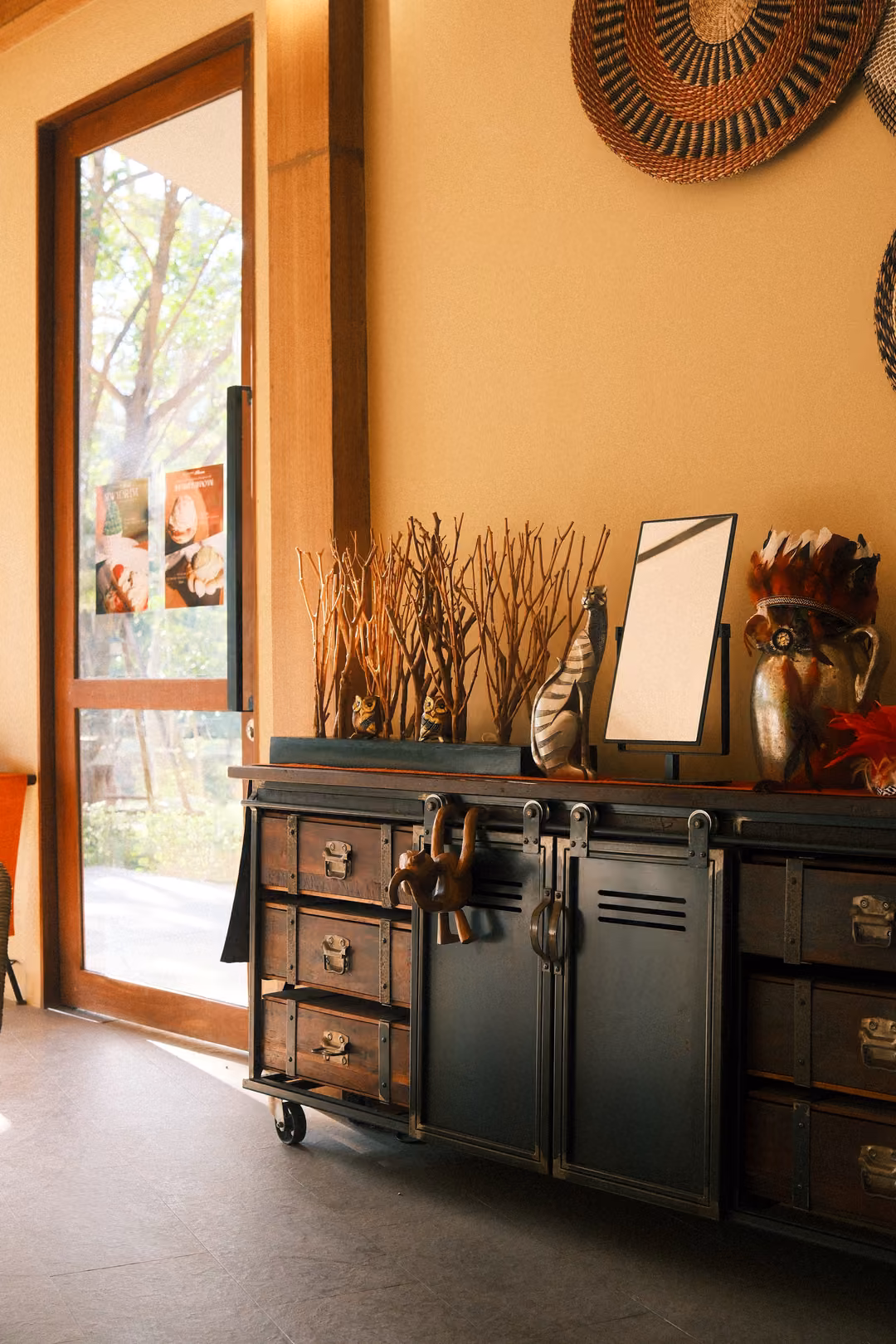 Dark cabinet with dried branches and a small mirror beside an open glass door, woven basket on a yellow wall