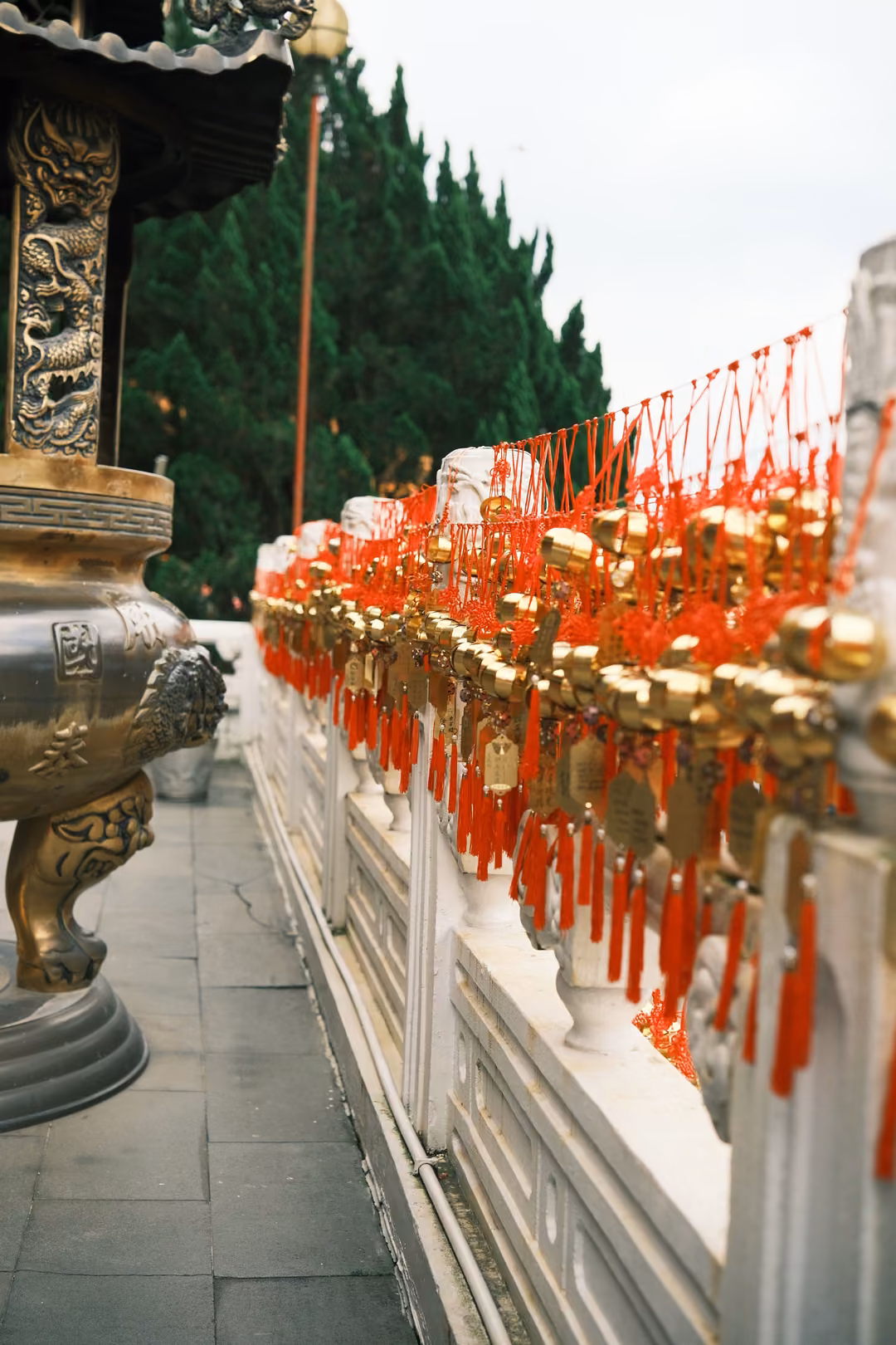 Row of red tassels and golden bells hanging along a white stone railing beside a bronze urn and tall pine trees