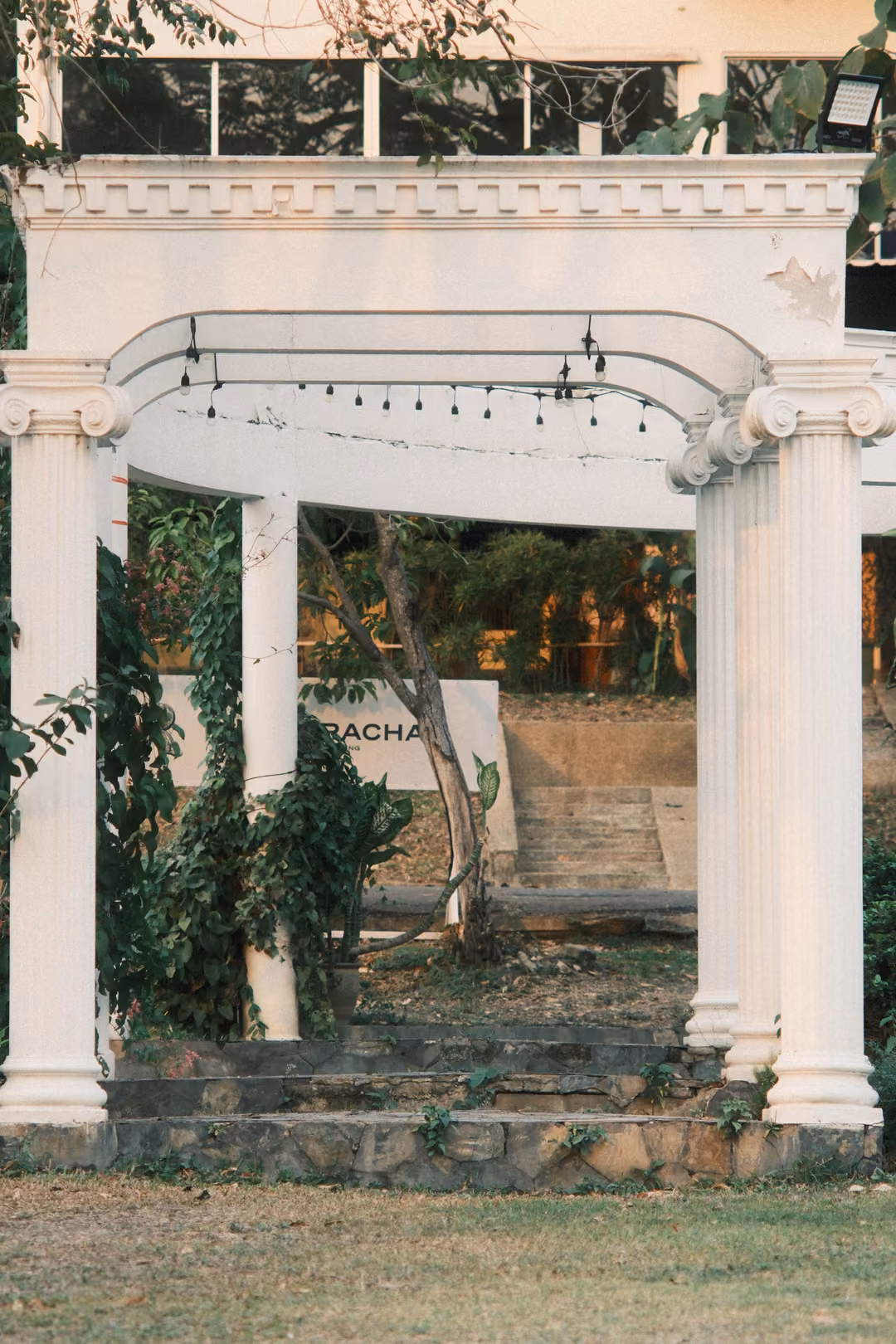 White arched colonnade with string lights, framing stone steps and garden beyond, in vertical format
