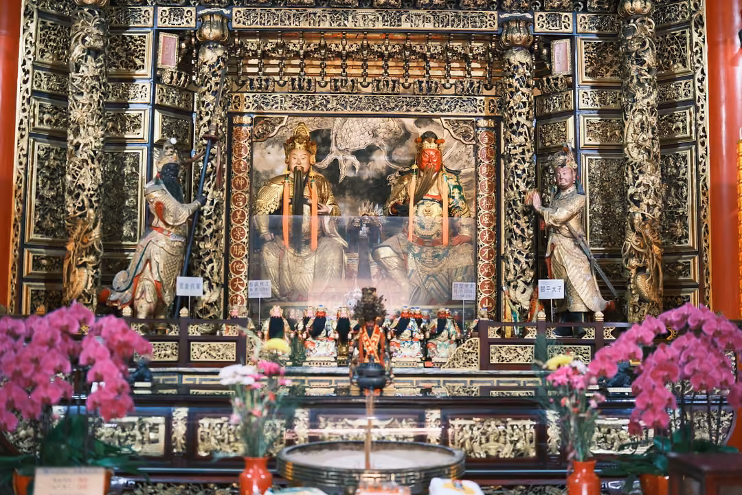 Wide view of a gilded altar with two seated figures, standing guardians and banks of magenta orchids