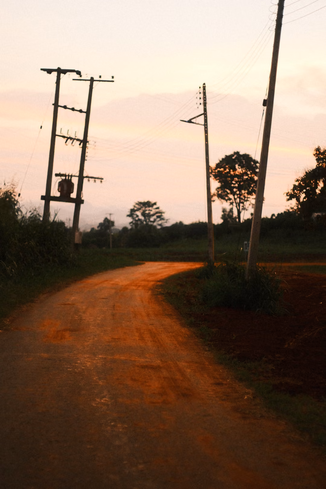 Red dirt road curving past power poles with a transformer, trees and pink evening sky