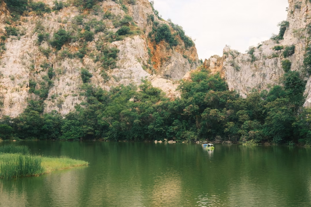 Limestone cliffs with dense trees reflected in a calm green lake, reeds on the left and a small pedal boat