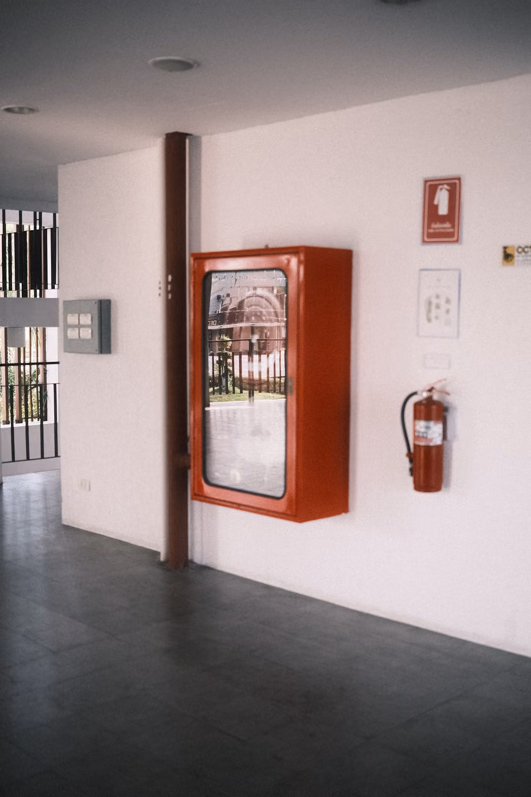 Red fire hose cabinet and extinguisher mounted on a white wall in a shaded hallway with grey floor tiles