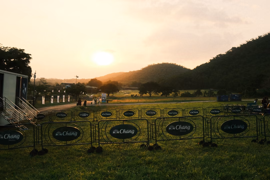 Sun setting over wooded hills behind a grass field lined with metal barriers bearing Chang logos.