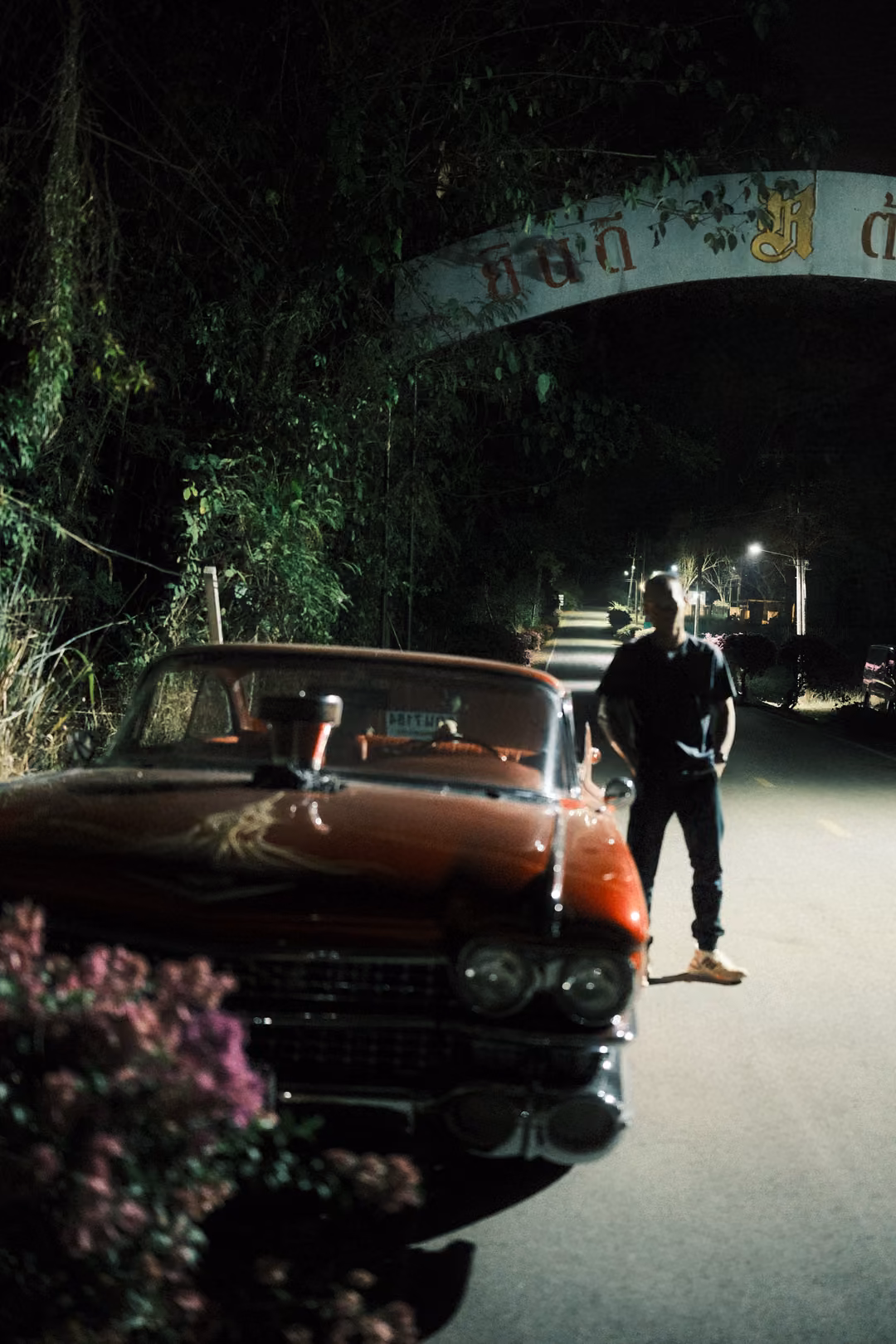 Man standing beside the front of a red vintage car on a dark road under an arched sign, flowers in foreground