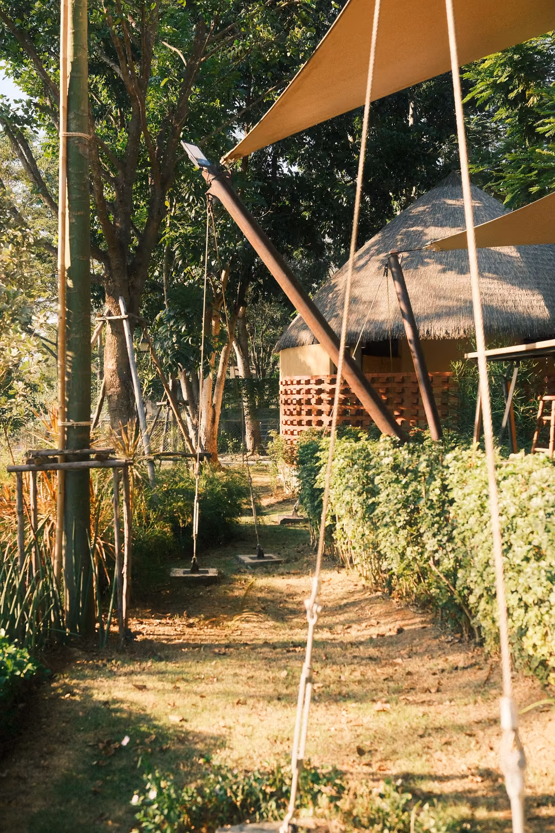Rope swings hanging from a canvas canopy over a grassy path, leading toward a thatched hut