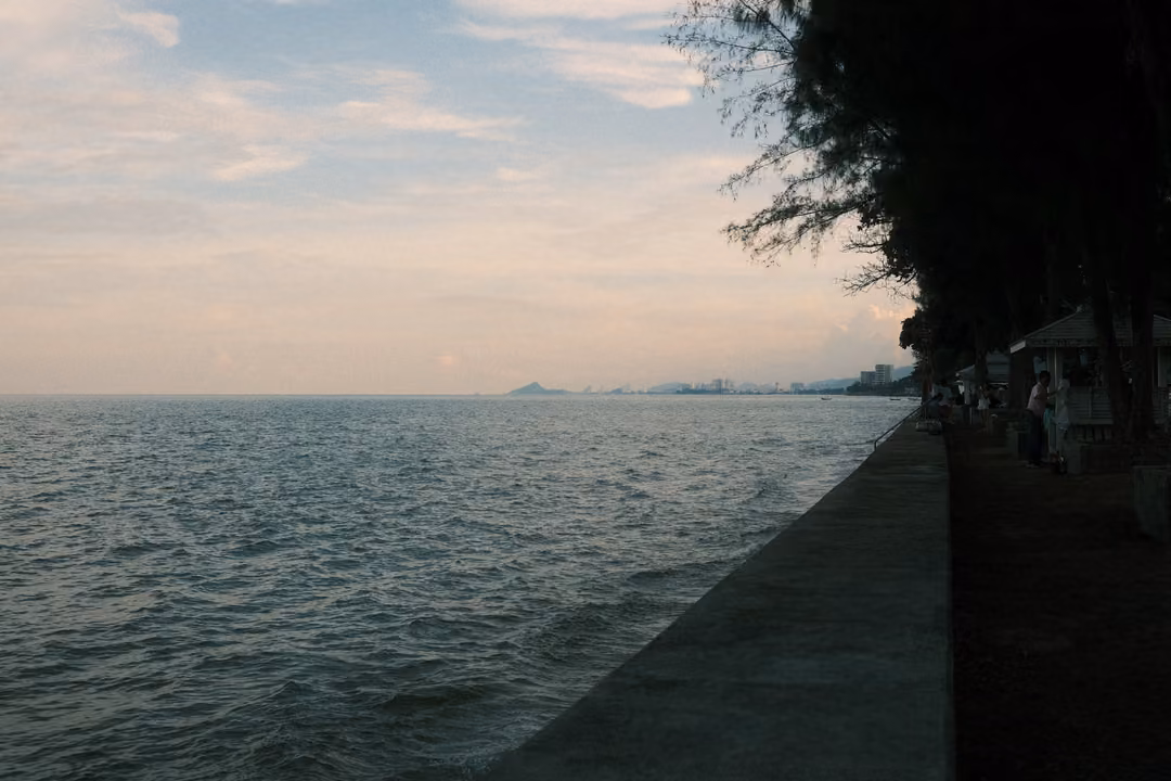 Seawall running along the water's edge toward distant hills and buildings, trees and lamps on the right