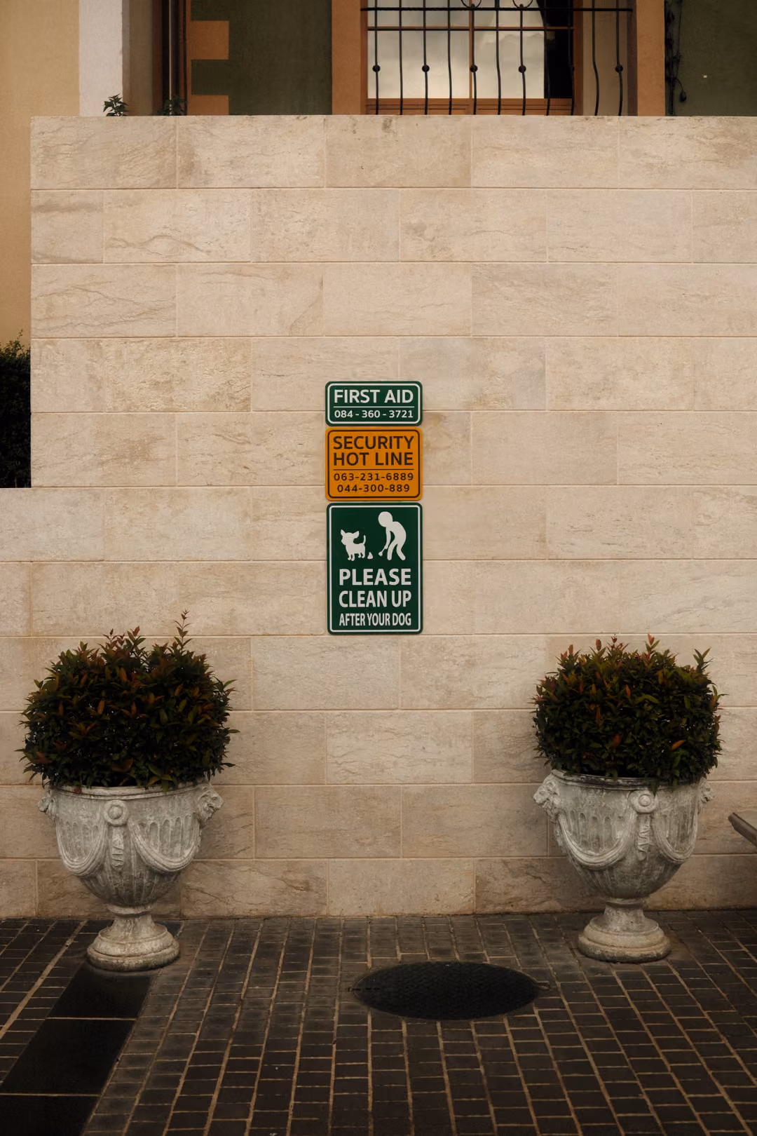 Pale stone wall with small First Aid, security and dog signs between two stone urns holding round shrubs