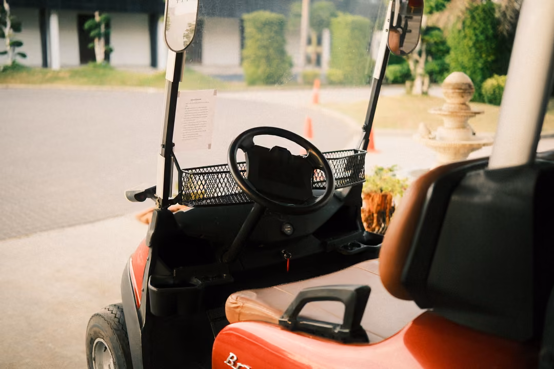 Close view from the seat of a red golf cart, its steering wheel and basket, a paved road and hedges beyond