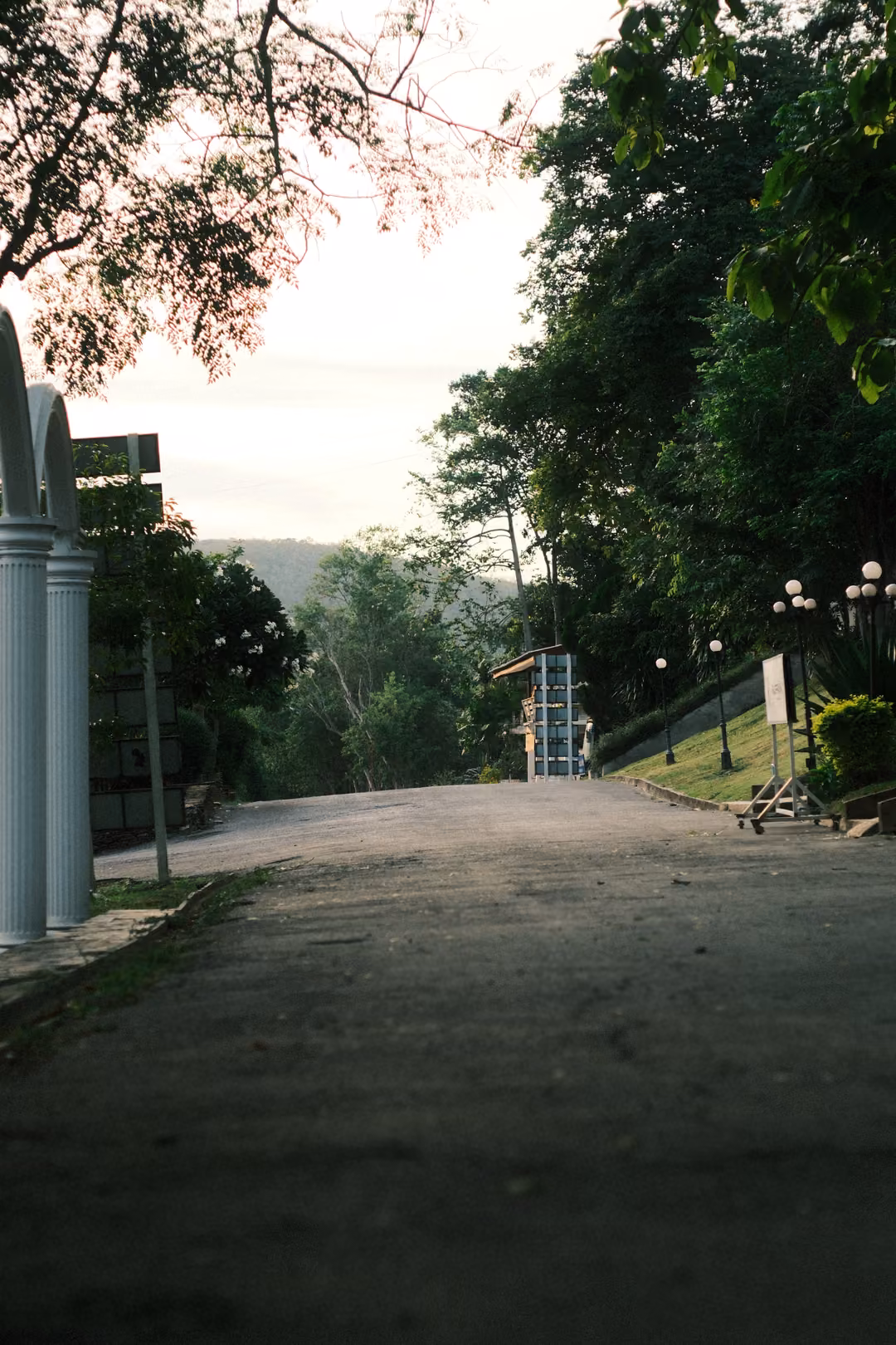 A road sloping away past white columns, ornate lamp posts and green trees, hills and a tower in the distance.