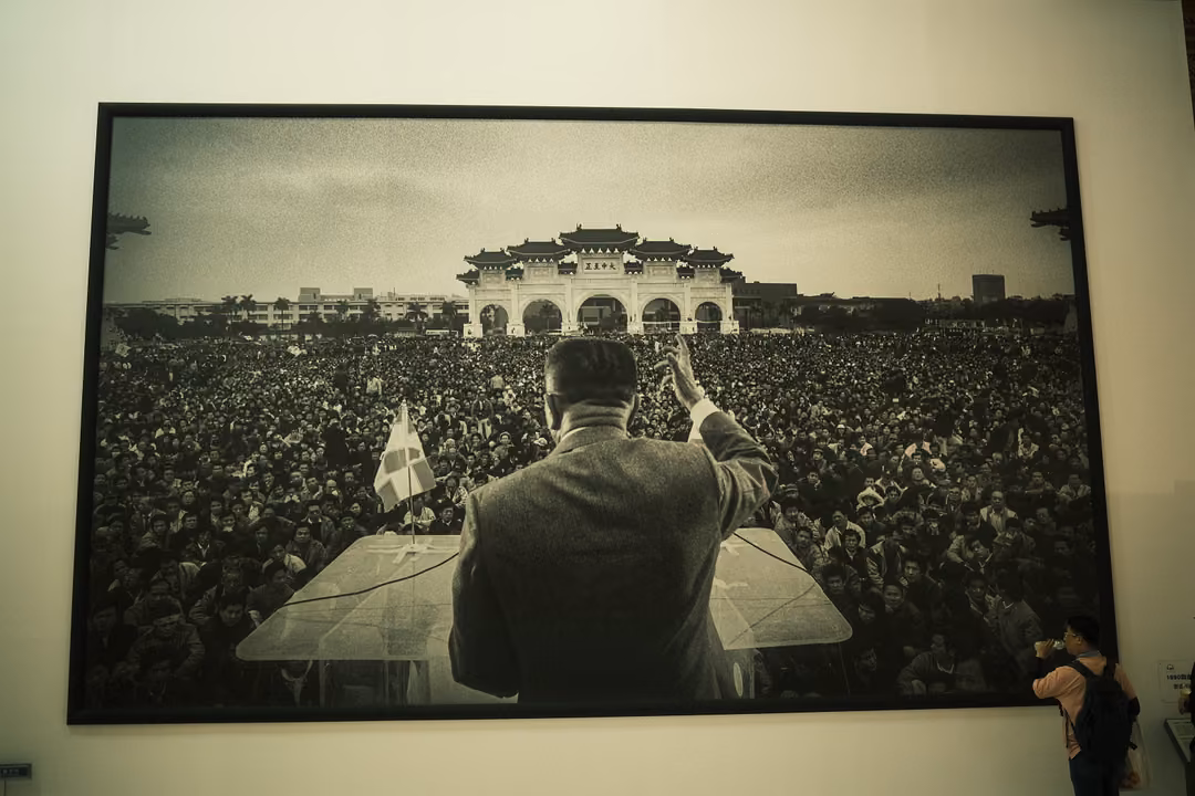 Large black-and-white photograph of a man waving to a vast crowd before an ornate gate, hung on a gallery wall