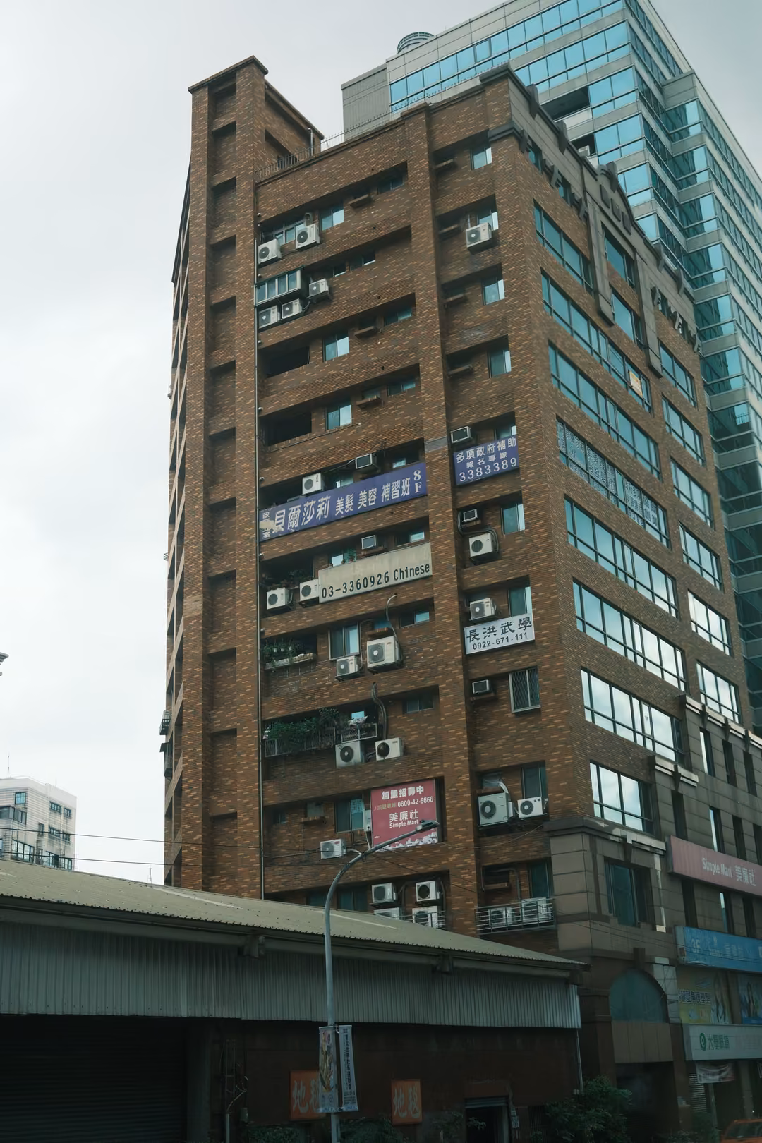 Tall brown brick apartment tower with air conditioners and signs beside a glass building, overcast sky