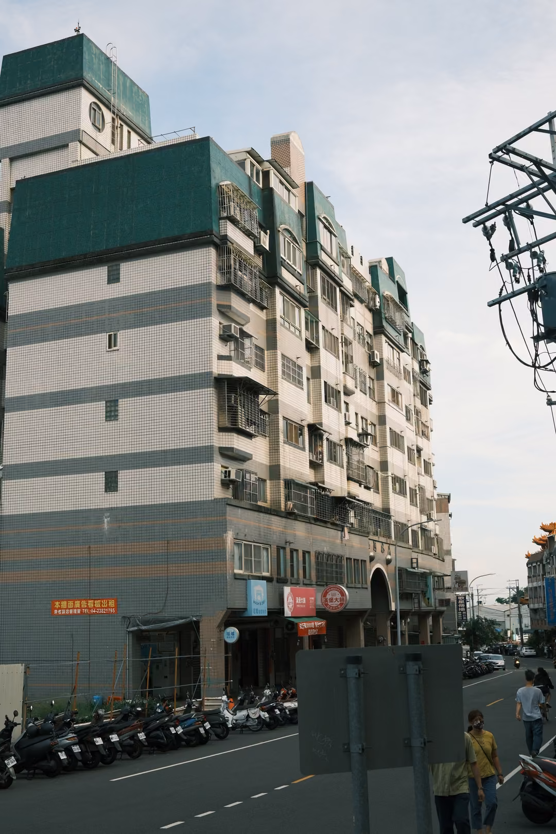 Apartment building with green tiled roof edge, utility pole, scooters lining the street and pedestrians