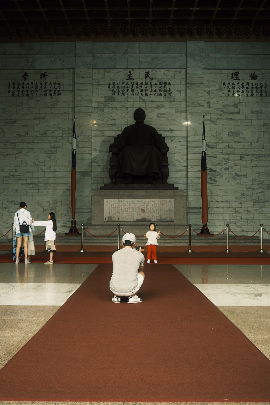 Wide hall with a long red carpet leading to a seated statue; a crouching man photographs two children