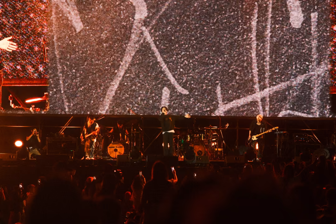 Band on stage below a dark screen with white scratchy lines, crowd with raised arms in red light.