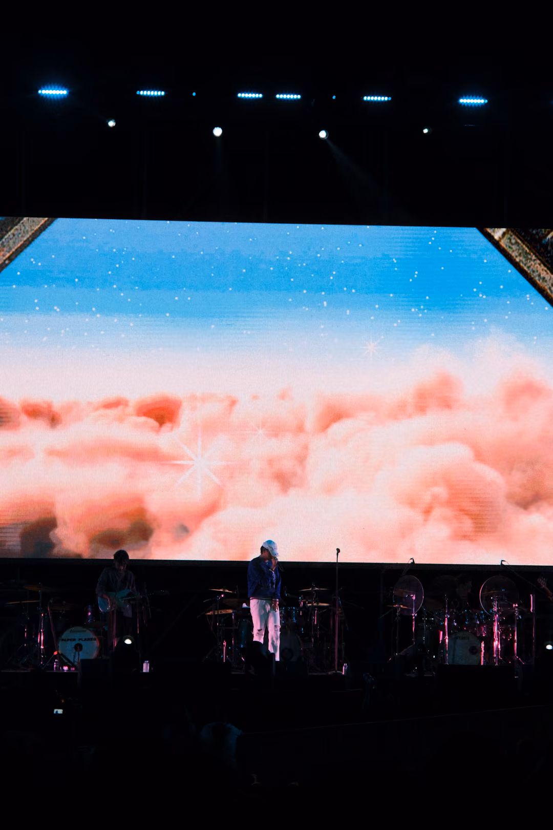 Vertical view of a screen showing a sky with a band of pink clouds below blue, the singer on stage in white trousers.