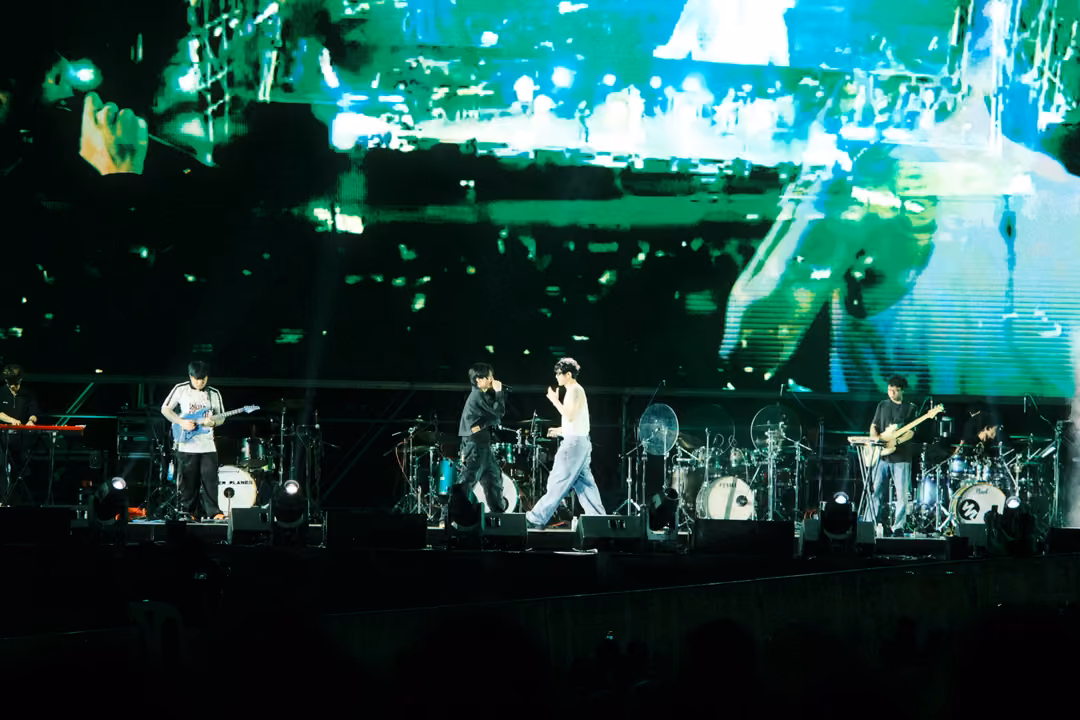 Band performing beneath a green screen showing a blurred live view of the stage rig and a singer's shoulder.
