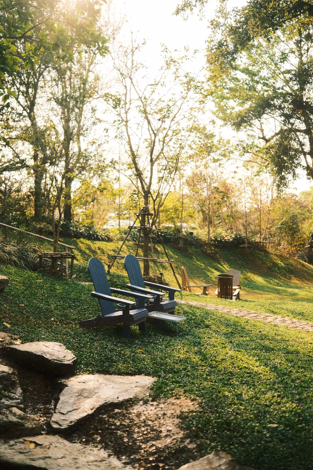 Two dark blue wooden lounge chairs on a sloping lawn with rocks in front and sun haze through trees