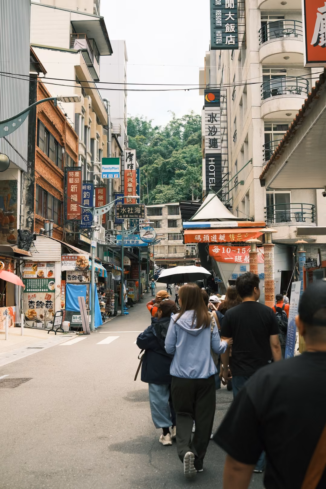 Busy narrow street with hanging shop signs and a HOTEL sign, people walking away, umbrellas and lanterns