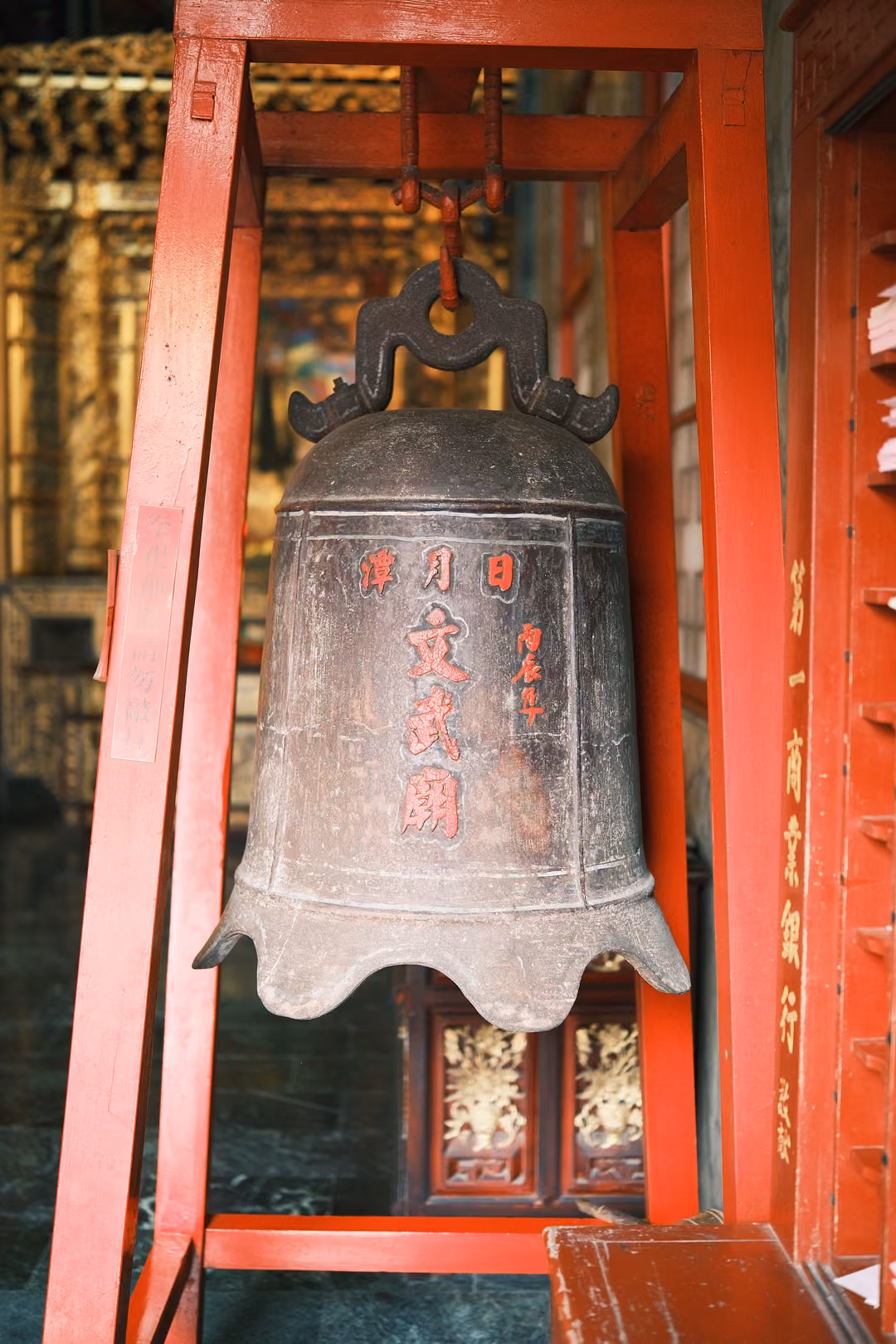 Closer upright view of the bronze bell with red characters in its red frame, gilded carvings behind