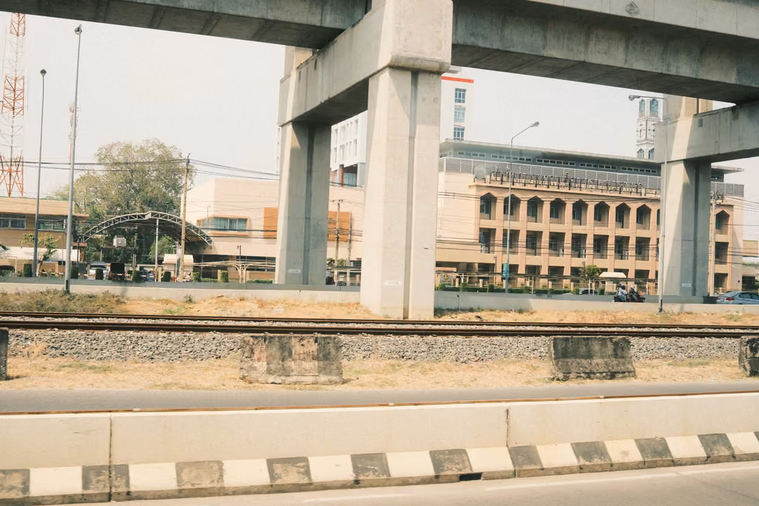 Concrete elevated rail pillars framing a cream building with arched windows, beyond railway tracks