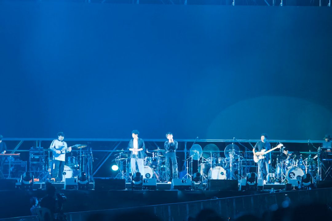 Band on stage in front of a deep blue backdrop, singers standing together at centre, crowd heads in shadow.