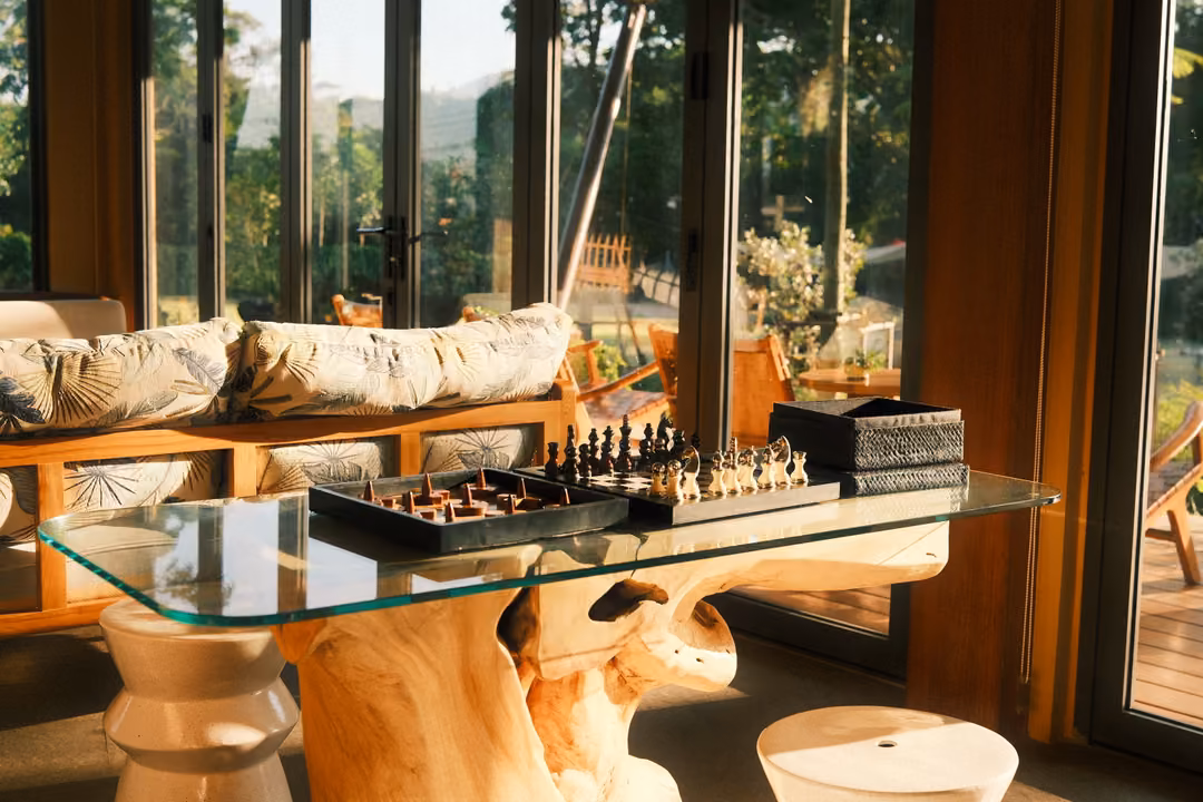 Glass table on a carved wooden base holding chess sets, white stools around it, sofa and windows behind