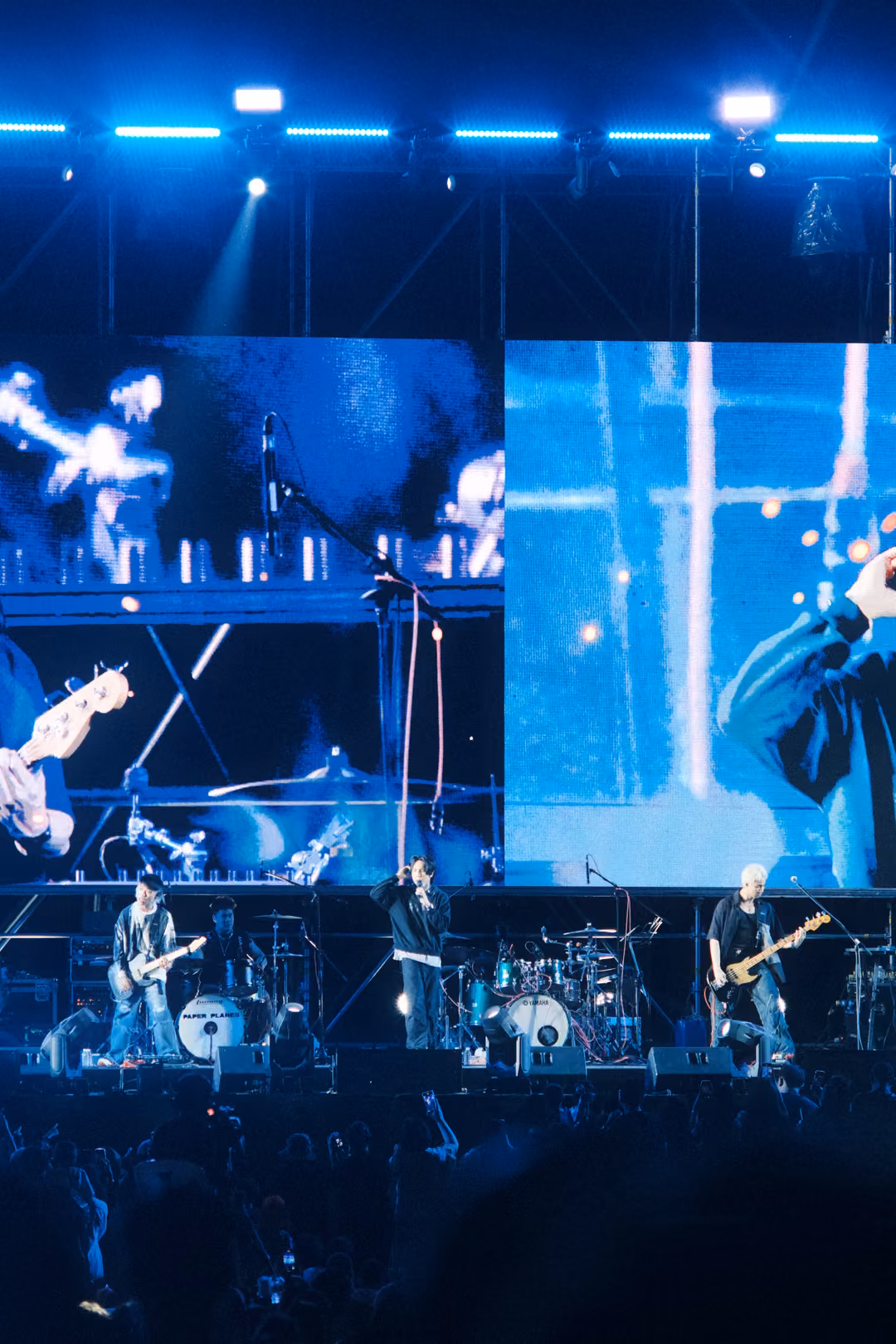 Vertical view of a stage under blue screen panels with a white cross shape of light, three musicians performing.