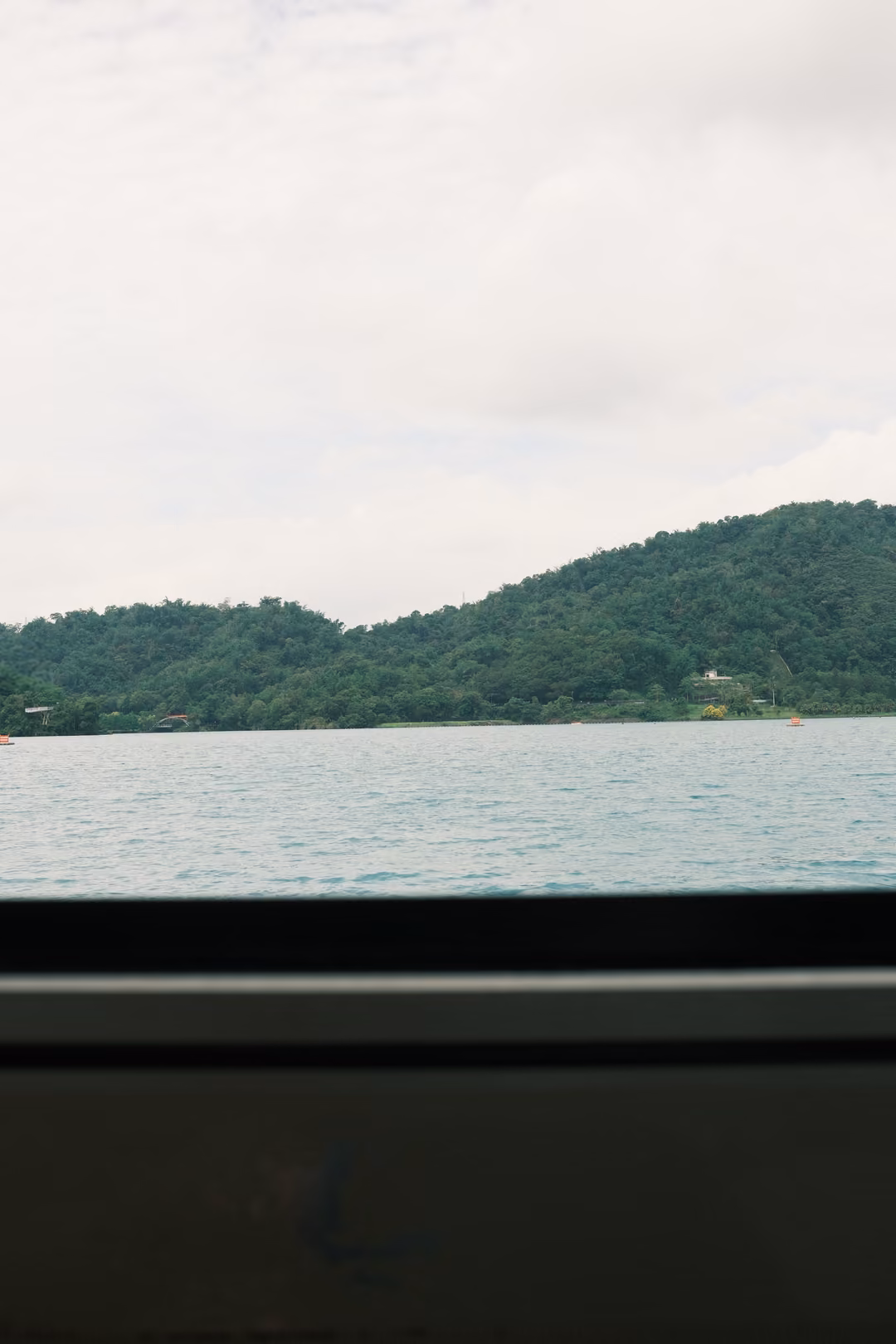 Vertical view of a wooded hill and lake framed by a boat window ledge, overcast sky above