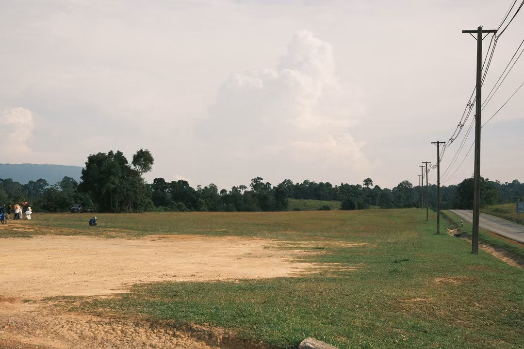 An open field with a patch of bare earth, power lines running to the horizon and a tall cloud above hills.