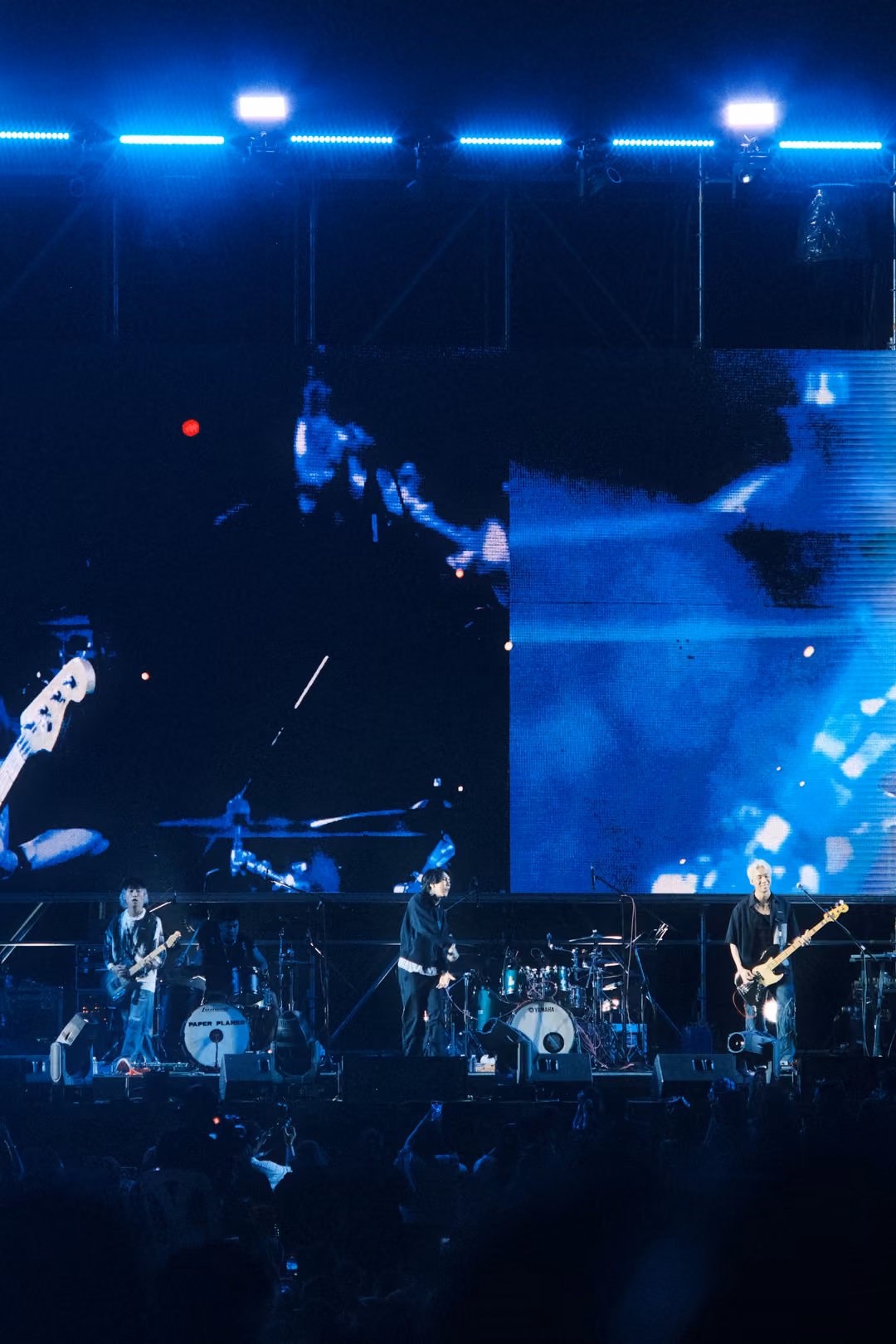 Vertical view of a dark stage with a blurred blue screen showing a guitar neck and light streaks, band below.