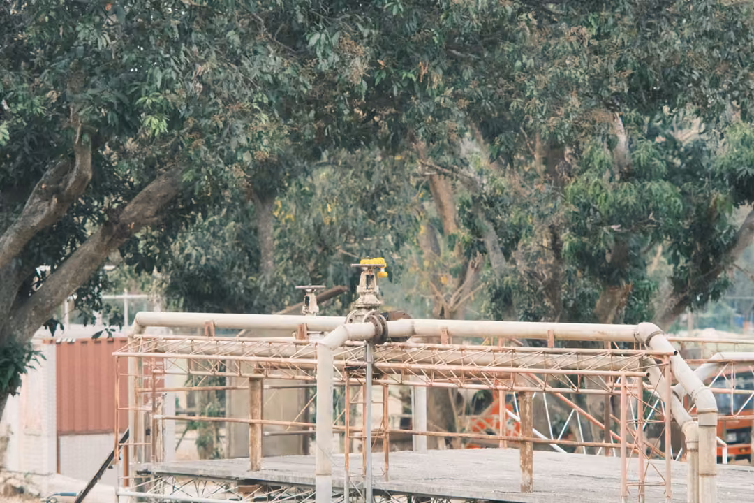 Rusty metal scaffold and pipe structure with a valve on top, under green trees beside a red wall