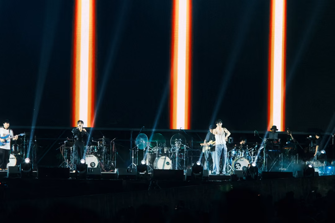 Band on stage below a black screen with three tall orange-and-white vertical light bars, beams of light in the haze.