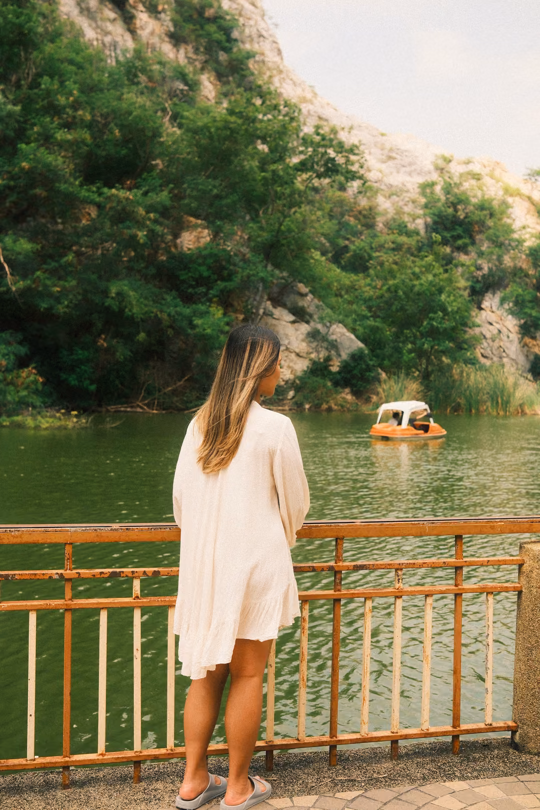 Woman in a white dress from behind at a lakeside railing, a small pedal boat crossing the water ahead of her